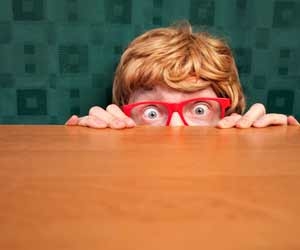 Scared boy in glasses hides behind a desk