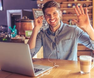 Young entrepreneur smiling and waving at camera while drinking coffee and working from laptop